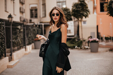 Fashionable pale brunette in long green dress, black jacket and sunglasses, standing on street...