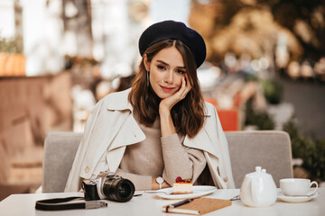 Pretty young brunette-haired girl with beret, trench coat and beige pullover, sitting at table with...