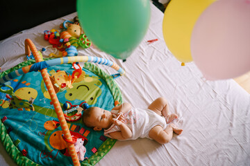 Baby lies on the bed next to toys and balloons