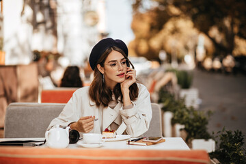 Thoughtful young brunette with red lips, beige trench coat and dark beret eating cheesecake and...