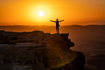 Man standing on a cliff with his arms open and looking at the sun rise over the Negev Desert in Israel - obrazy, fototapety, plakaty