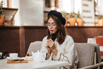Charming young dark-haired lady in beret, glasses and beige trench coat having rest at sunny city cafe terrace, eating cheesecake with tea, and looking down