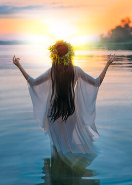 Fantasy woman standing in water hands raised to sky, praying to sun. Slovenian girl in herbal wreath on head, long hair white dress, pagan holiday of Ivan Kupala. Nature blue river sunset. Back view.