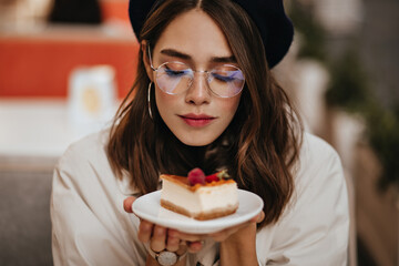 Pretty young girl with dark wavy hairstyle, modern makeup, stylish earrings and beige trench coat sitting at city cafe terrace and holding piece of cheesecake
