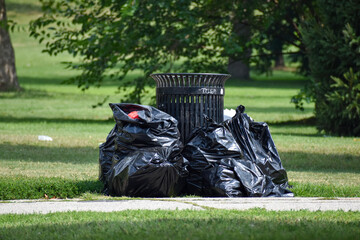 Many black trash bags piled against trash can in a beautiful  public park with lots of green grass...