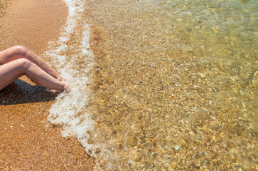 View of female sitting on sand beach with legs in oncoming wave. Greece. 