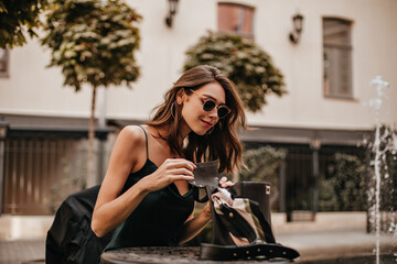 Charming young lady with brunette wavy hair, trendy sunglasses, green silk dress sitting outdoors...