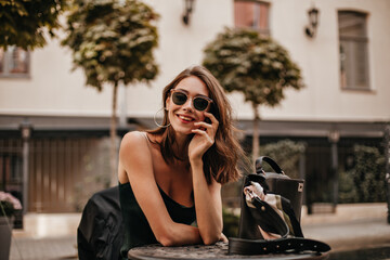 Engaging young dark-haired girl with red lips, modern sunglasses and silk slip dress, smiling and posing outdoors. Light buildings and trees background