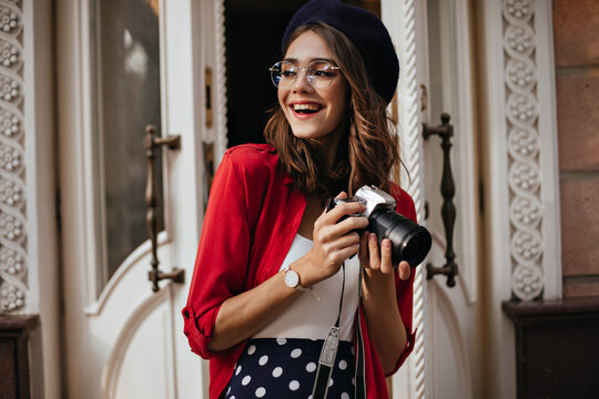 Pretty young Parisienne with red lips and brown hair, in classic outfit, beret and glasses making photo, smiling and looking away against vintage door background