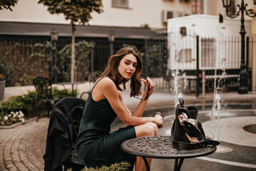 Elegant young brunette with well-built body in green silk dress, sitting at table and holding sunglasses. City cafe terrace during daytime