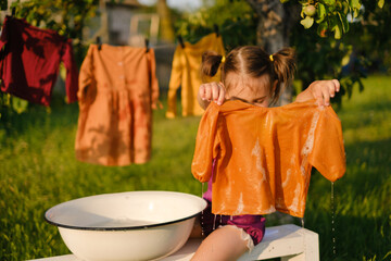 The child holds a shirt wet after washing in his hands. Hanging laundry is drying in the...