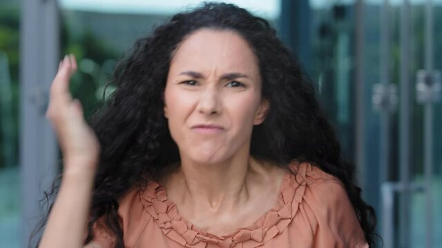 Female portrait of young Hispanic woman with curly long hair stands outdoors shouts angry waving hands. Annoyed in rage mad girl quarrel says bad words argument emotional abuse aggressive shouting