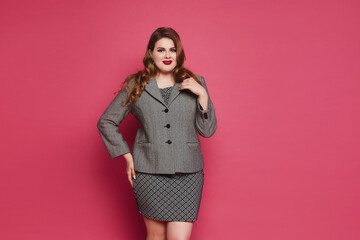 A young plump woman with bright makeup and red full lips wearing office clothes posing on the pink background. Plus-size model girl in grey dress and suit coat over pink background