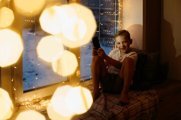 portrait of a boy with a phone in his hands at night at home. Child on the windowsill at christmas evening