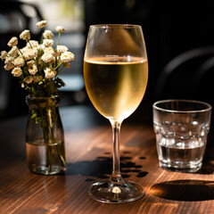 A glass of cold white wine back-lit by sunlight and glass of water on a dark brown wood table decorated with small white flowers. Serving lunch at winery. Refreshing drink on a summer day