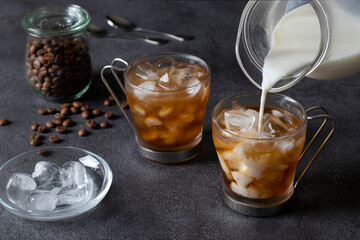 Two cups of cold coffee with ice cubes, into which milk is being poured from a jug. Dark background. Close-up.