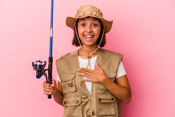Young mixed race fisherwoman holding a rod isolated on pink background laughs out loudly keeping hand on chest.