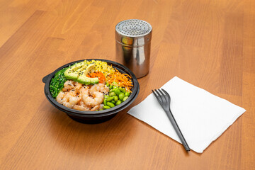 Poke bowl Hawaiian edamame beans, cooked shrimp, fish roe, chopped mango, grated carrot, ripe avocado, wakame seaweed salad in black home delivery container