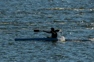 Person in the cockpit of a kayak propelling the craft with a double bladed paddle  racing across a lake in blue water, a  bright sunlight reflecting off the water. The wake of other racers can be seen