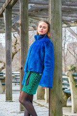 Dressing in a blue faux fur plus size jacket, patterned dress, a pretty girl is leaning against  a pillar of a wooden frame structure in winter, sad, looking forward..