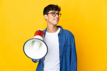 Young man over isolated yellow background holding a megaphone and looking up while smiling