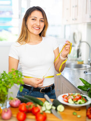 Positive colombian housewife measures waist before eating vegetable salad at kitchen