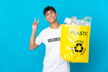 holding a bag full of plastic bottles to recycle over blue background smiling and showing victory sign