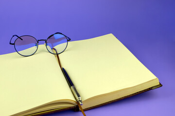 Open notebook and round glasses. Stylish glasses, notepad and pen, close-up. Blank pages of a notebook and glasses on a purple background