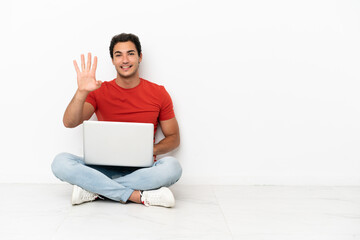 Caucasian handsome man with a laptop sitting on the floor happy and counting four with fingers