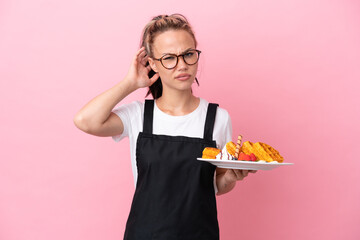 Restaurant waiter Russian girl holding waffles isolated on pink background having doubts