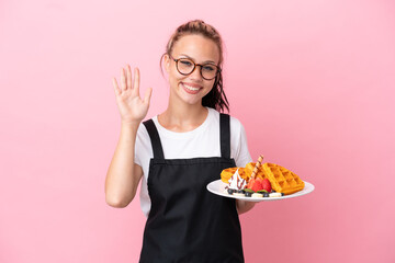 Restaurant waiter Russian girl holding waffles isolated on pink background saluting with hand with happy expression