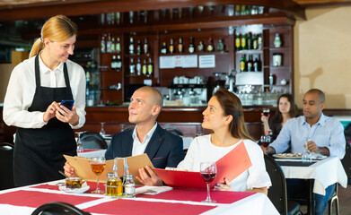 Young smiling waitress consulting couple of guests choosing drinks and meals in cozy pizzeria