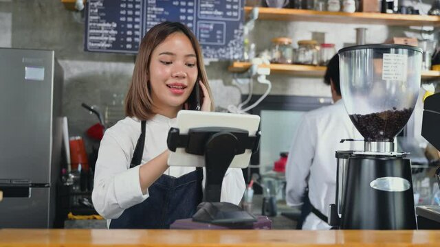 Owner coffee shop and online order concept. Asian young woman barista in apron talking on mobile phone and using tablet to customer in coffee shop. Small business cafe restaurant retail food and drink