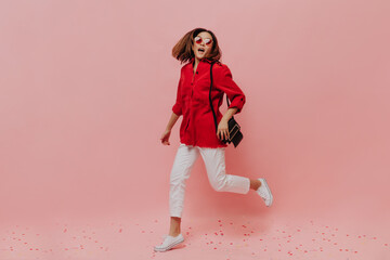 Short-haired woman in red oversized shirt and white pants run on pink background. Lady in sunglasses holds handbag and moves on isolated.