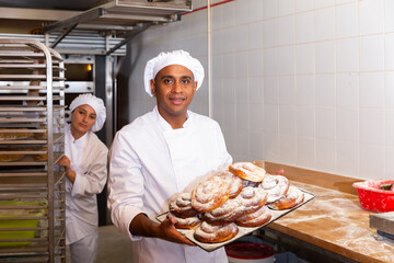 Smiling latin american bakery worker carrying tray with fresh baked ensaimadas sprinkled with powdered sugar