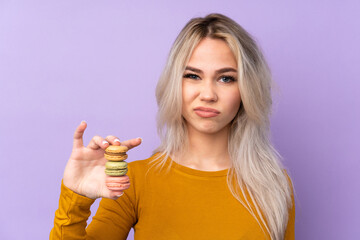Teenager girl over isolated purple background holding colorful French macarons and with sad expression