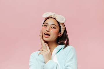 Pretty young woman in soft blue pajamas and beige headband touches her face, looks into camera and poses with cosmetic eyepatches.