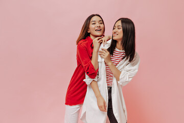 Young attractive women have fun on isolated pink background. Cute lady in red shirt and white pants and her friend in stylish outfit smile.