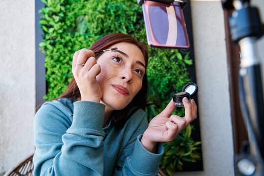 young caucasian woman with red hair doing her makeup in front of a tripod with circular light and her cell phone. female influencer blogger live in front of her smartphone. social media concept