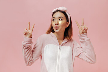 Charming young woman in pajamas whistles and shows peace signs on pink isolated background.