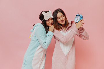 Cute short-haired girls in soft pajamas pose on pink background. Young girls in kigurumi holds blue alarm clock on isolated.