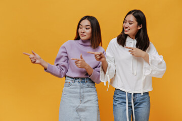 Charming Asian women in denim outfits point to left at place for text on isolated. Brunette girls...