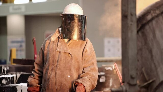 An industrial foundry worker in protective clothing supervising the careful ladle pouring of molten steel into the casting mold.