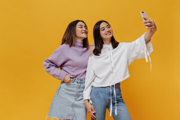 Tanned brunette Asian women in denim outfits and sweatshirts take selfie on isolated. Pretty cool...