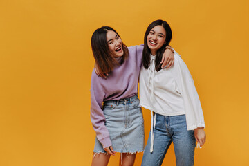 Joyful happy emotional girls laugh on orange background. Pretty Asian woman in denim skirt and purple sweater hugs friends on isolated.