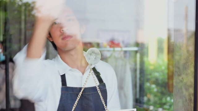 View through door glass of Asian young man barista in apron using spray and cloth with cleaning door glass before open in coffee shop. Male waiter wash wipe clean glass by spray alcohol in restaurant.