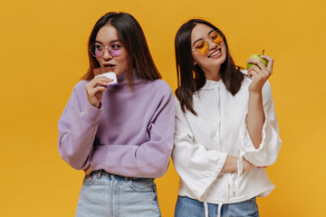 Pretty girl in purple sweater bites milk chocolate bar. Young woman in white hoodie looks at green apple and smiles. Portrait of charming Asian women pose on orange background.