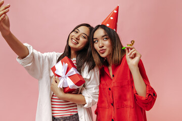 Tanned brunette Asian women take selfie and celebrate birthday on pink background. Cute girl in white shirt poses with red gift box. Charming lady in party hat holds party horn.