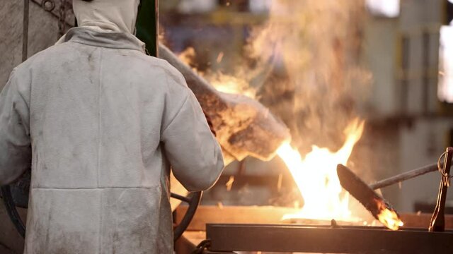 A foundry worker pours hot, firey, liquid metal, with dramatic glowing white sparks and smoke, into a casting mold at an engineering factory.