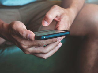 Close up hand man holding smartphone. A young person, man in casual grey shirt and shorts sitting and interacting with technology at home. Relaxation male using mobile phone.